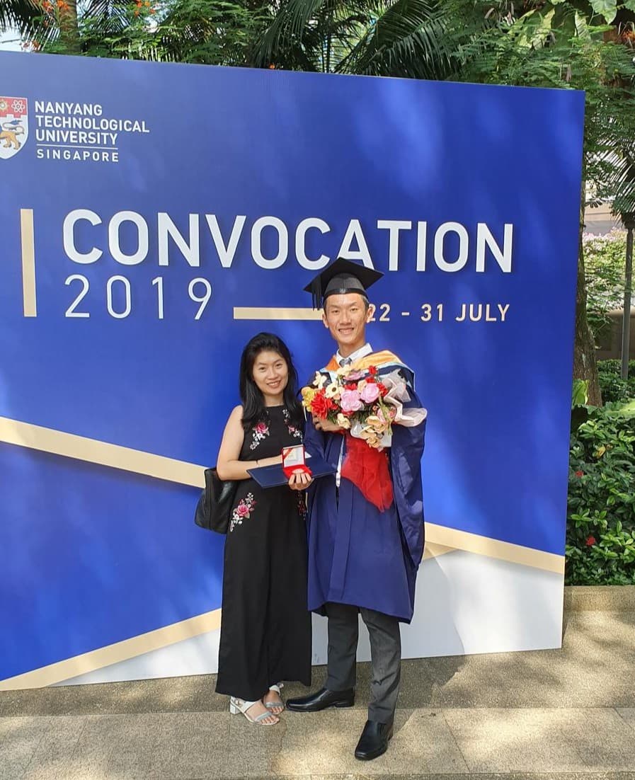 A graduate in an NTU gown holding a bouquet beside a woman, in front of the NTU Convocation 2019 backdrop